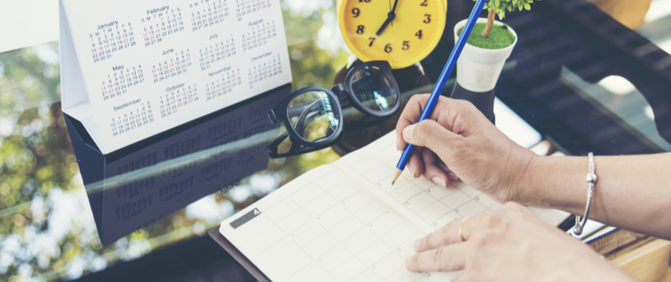 Person writing in planner with a blue pencil on a desk filled with a calendar, yellow clock, plant, and glasses.